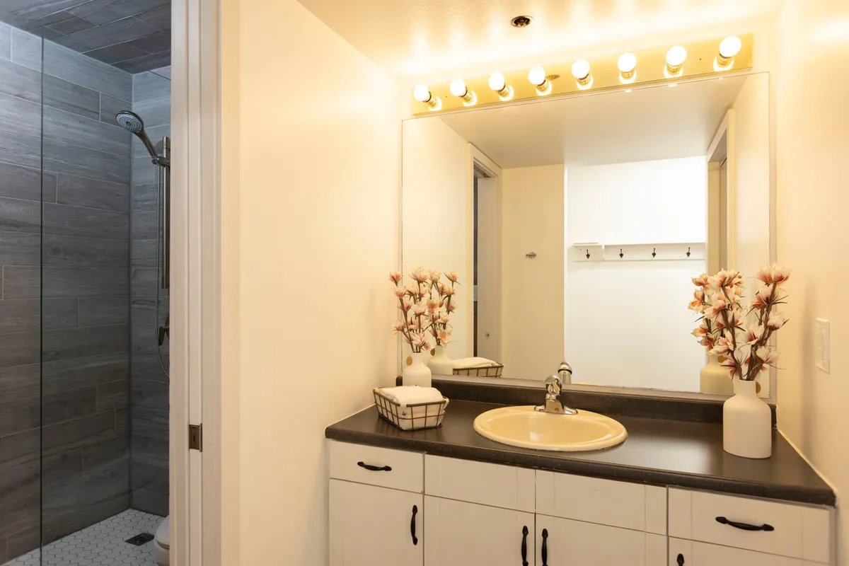 A serene bathroom scene featuring a lighted mirror, modern sink, decorative flowers, and a glimpse of the shower area. The décor is tranquil and soothing, with thoughtful details that contribute to a restful environment. The mood is calm, perfect for winding down or preparing for a restorative night's sleep.