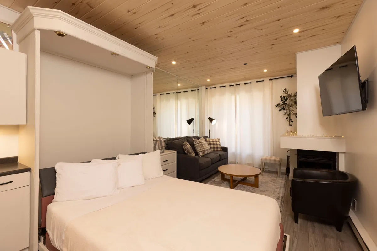The hero image shows a cozy, modern studio interior featuring a plush sofa, a Murphy bed, a wood-burning fireplace, and a coffee table arranged for comfort and connection. The warm glow of the fire and inviting seating area create a perfect setting for intimate movie nights or relaxing mornings with coffee.
