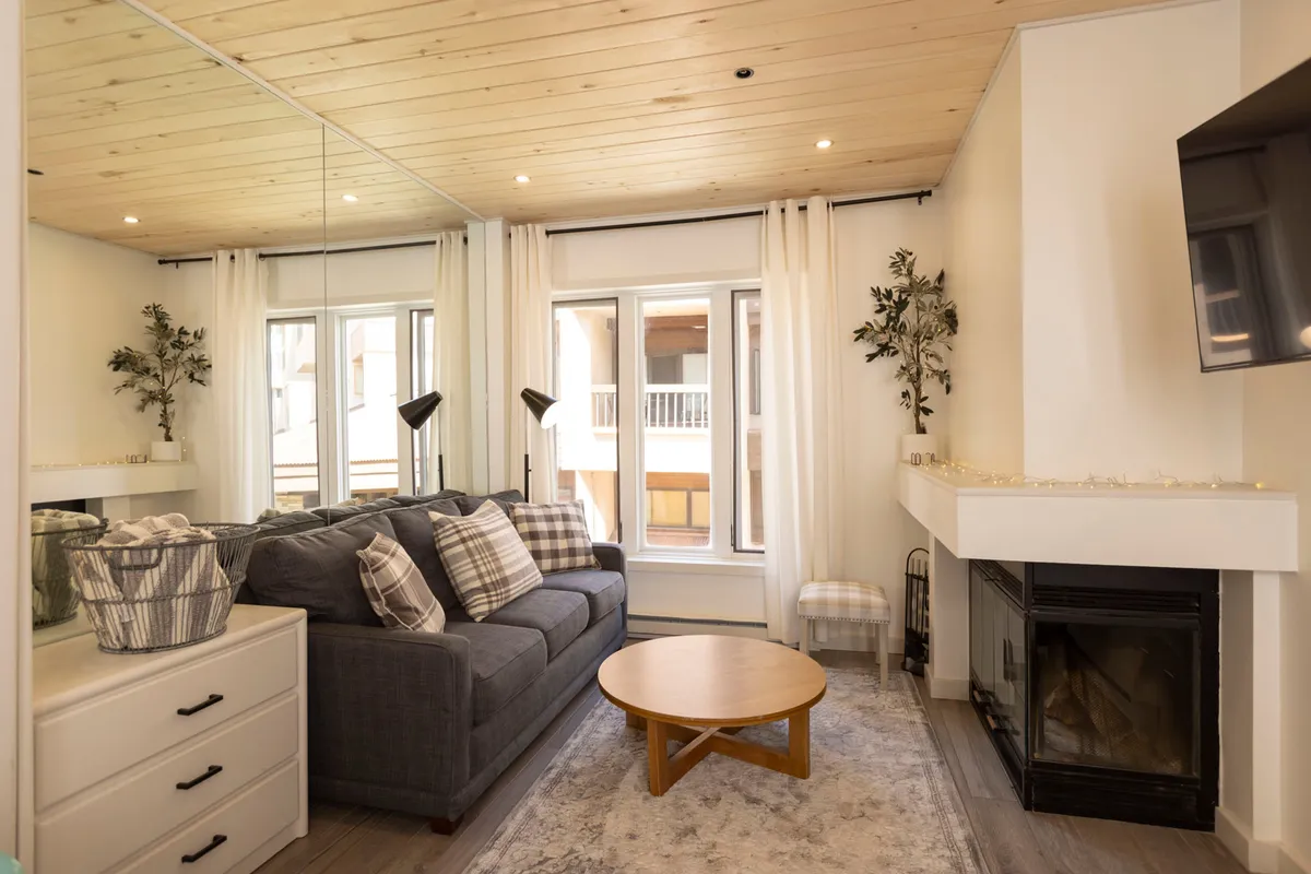 A warmly lit living area featuring a plush sofa and coffee table positioned around a striking wood-burning fireplace, with large windows letting in natural sunlight. The space exudes comfort and is arranged to invite conversation, relaxation, and social gatherings.