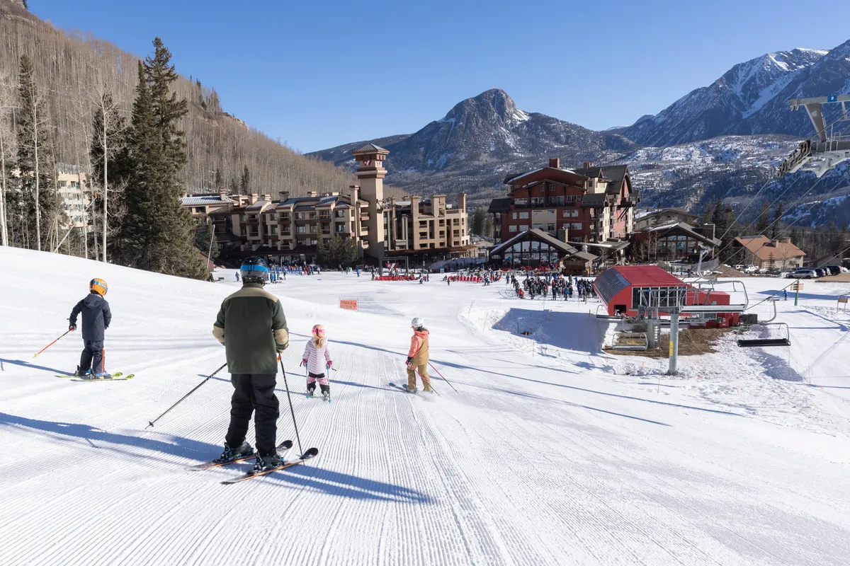 This vibrant exterior image captures skiers descending snowy slopes with the resort and ski lift visible in the background, all framed by the surrounding mountains. The scene is lively, showing a mix of individuals and families enjoying winter sports in direct proximity to the condo building.