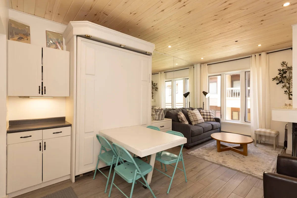 A welcoming studio interior with a cozy sofa positioned near large windows that flood the space with natural light. The wooden ceiling adds warmth and enhances the tranquil atmosphere. A folding table offers a multi-functional area for relaxed dining or quiet activities. The overall ambiance is comfortable and inviting, perfect for unwinding.