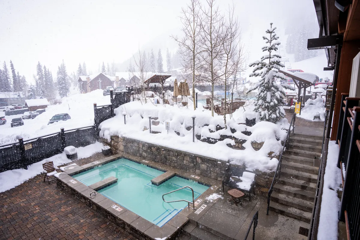 A steaming outdoor hot tub set against a snow-covered landscape, with trees nearby and steps leading up. The setting is tranquil, evoking relaxation, and hints at stargazing or snow-watching while soaking in the tub.