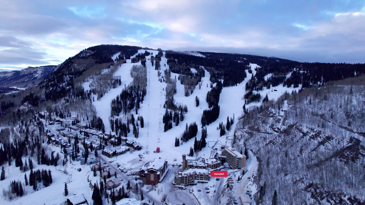 A panoramic daytime shot showcasing the dramatic ski slopes, mountain backdrop, and residential resort buildings. Ski lifts traverse the frame, emphasizing the area's adventure and accessibility to mountain experiences.