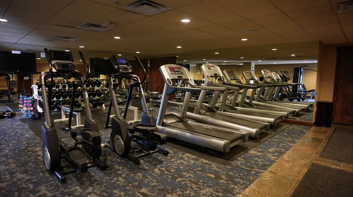 A modern fitness center interior featuring elliptical machines, treadmills, a dumbbell rack, and mirrored walls. The space appears energetic, bright, and motivates an active lifestyle.