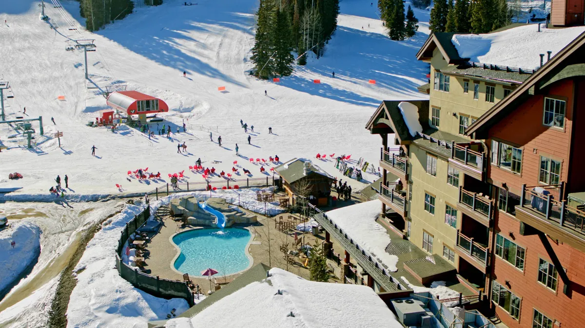 A lively outdoor scene that features a heated pool nestled beside lodge buildings, with visible ski slopes and skiers in the background. The atmosphere is festive, blending relaxation with the energy of the resort.